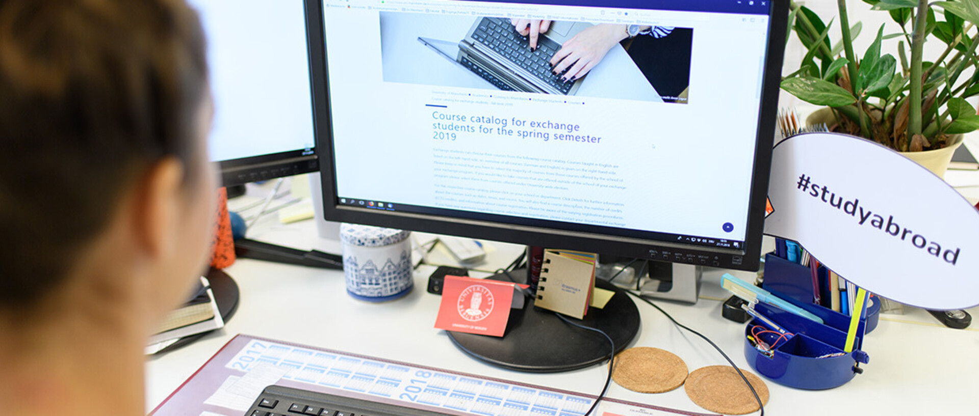 Eine Person sitzt vor einem Computerbildschirm und schaut sich eine Webseite zum Thema 'Studieren im Ausland' an. Auf dem Schreibtisch befinden sich diverse Gegenstände wie Stifte, eine Tasse und ein Schild mit der Aufschrift '#studyabroad'.