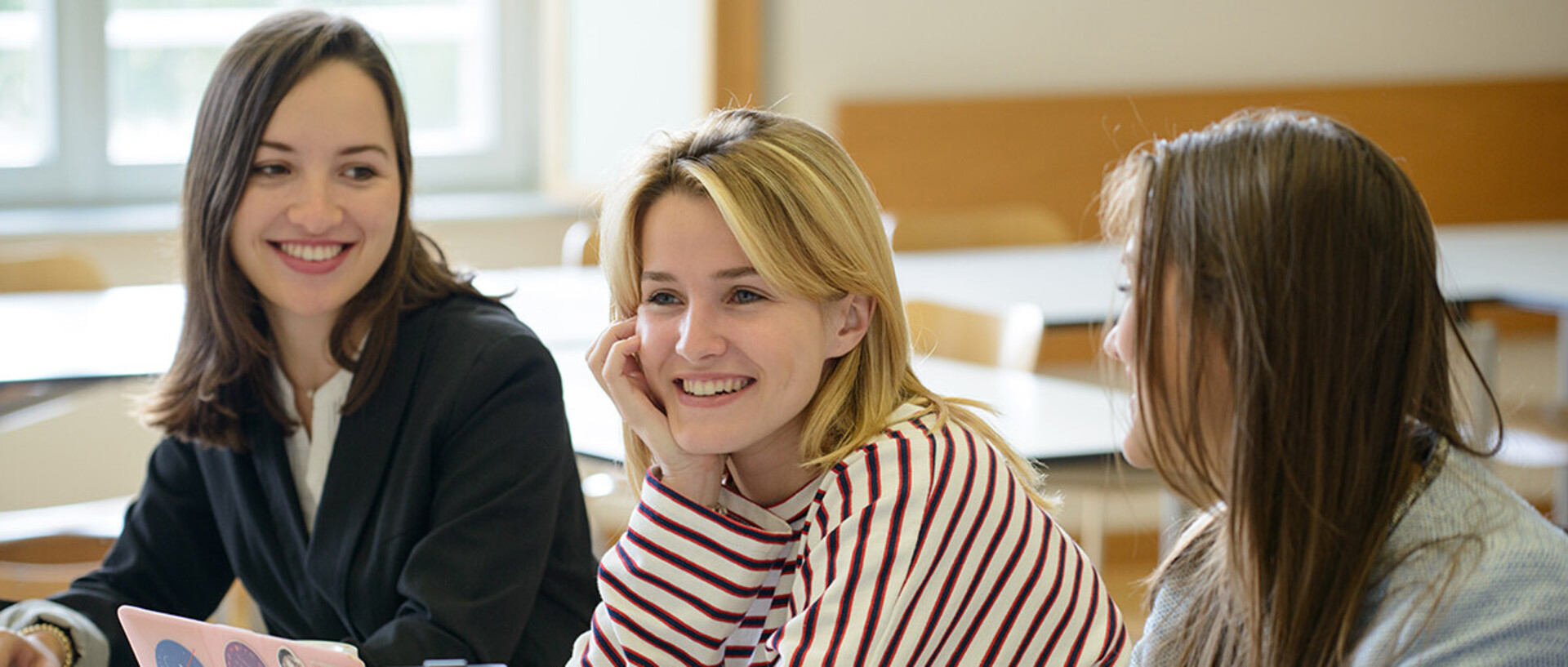 Three female students sit laughing in a seminar room.