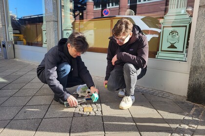 Zwei Mitglieder des Otto-Selz-Instituts, links Professor Alpers und rechts Sven Cornelisse, knien auf dem Gehweg neben dem Stolperstein von Otto Selz, den sie gerade reinigen. Professor Alpers hält eine Flasche und einen Putzlappen, während Sven Cornelisse aufmerksam zusieht. Im Hintergrund spiegelt sich die urbane Umgebung in den Fensterscheiben eines Gebäudes.