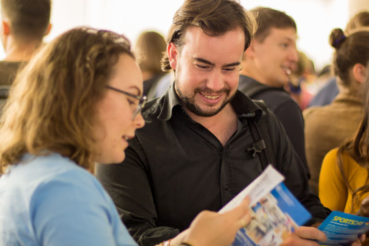 Drei Personen schauen sich jeweils einen Flyer an. Im Hintergrund sind weitere Menschen.