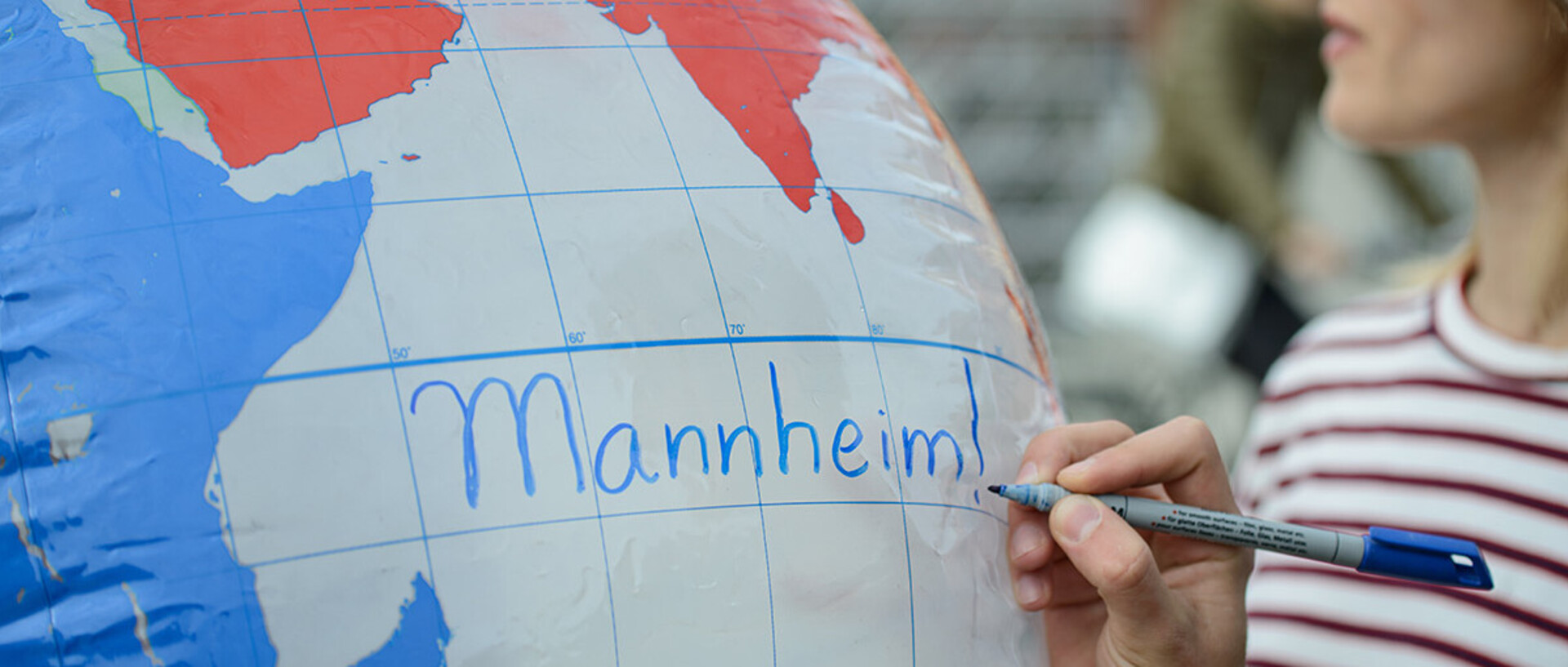 Eine Hand schreibt "Mannheim!" in blauer Schrift auf eine aufgeblasene Weltkugel.