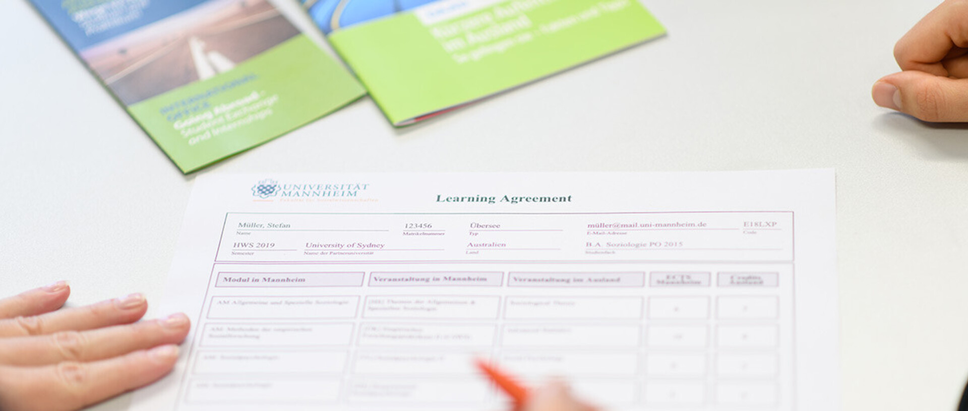 Zwei Personen füllen gemeinsam ein 'Learning Agreement'-Formular aus, wobei eine Person mit einem Stift auf das Dokument zeigt. Im Hintergrund liegen Informationsbroschüren in Grün- und Blautönen auf dem Tisch.