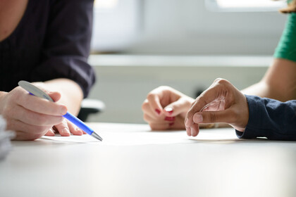 Zwei Studierende werden beraten, dabei sind die Hände und der Stift der Mitarbeiterin, der auf ein Papier zeigt, im Fokus.
