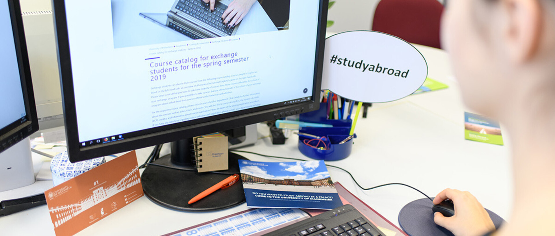Eine Person sitzt vor einem Computerbildschirm und schaut sich eine Webseite zum Thema 'Studieren im Ausland' an. Auf dem Schreibtisch befinden sich diverse Gegenstände wie Stifte, eine Tasse und ein Schild mit der Aufschrift '#studyabroad'.